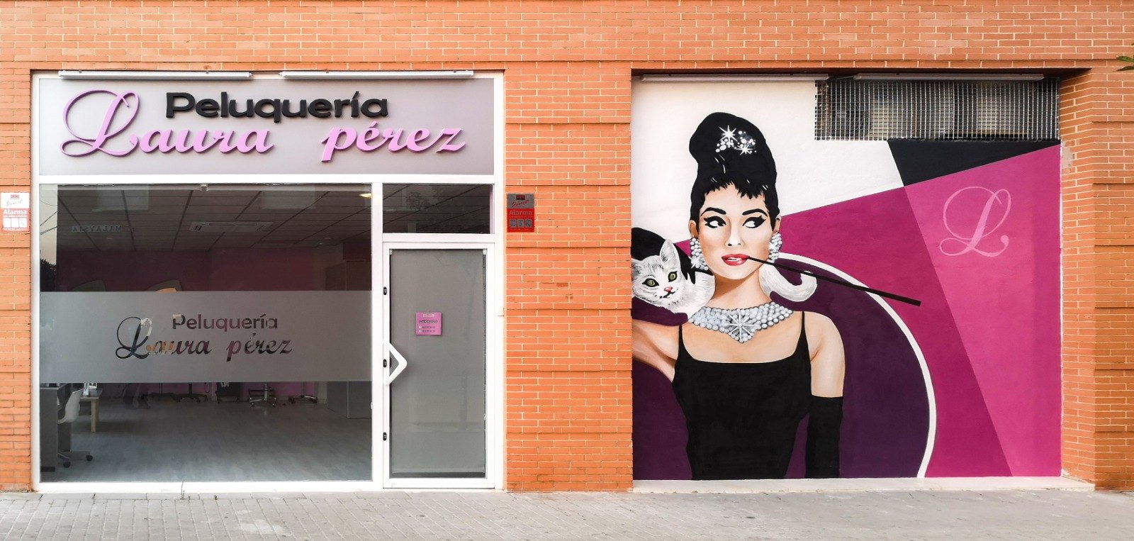 Fachada de ladrillo de la Peluquería Laura Pérez, con el rótulo en letras lilas y un mural de una mujer con moño y collar sobre fondo magenta